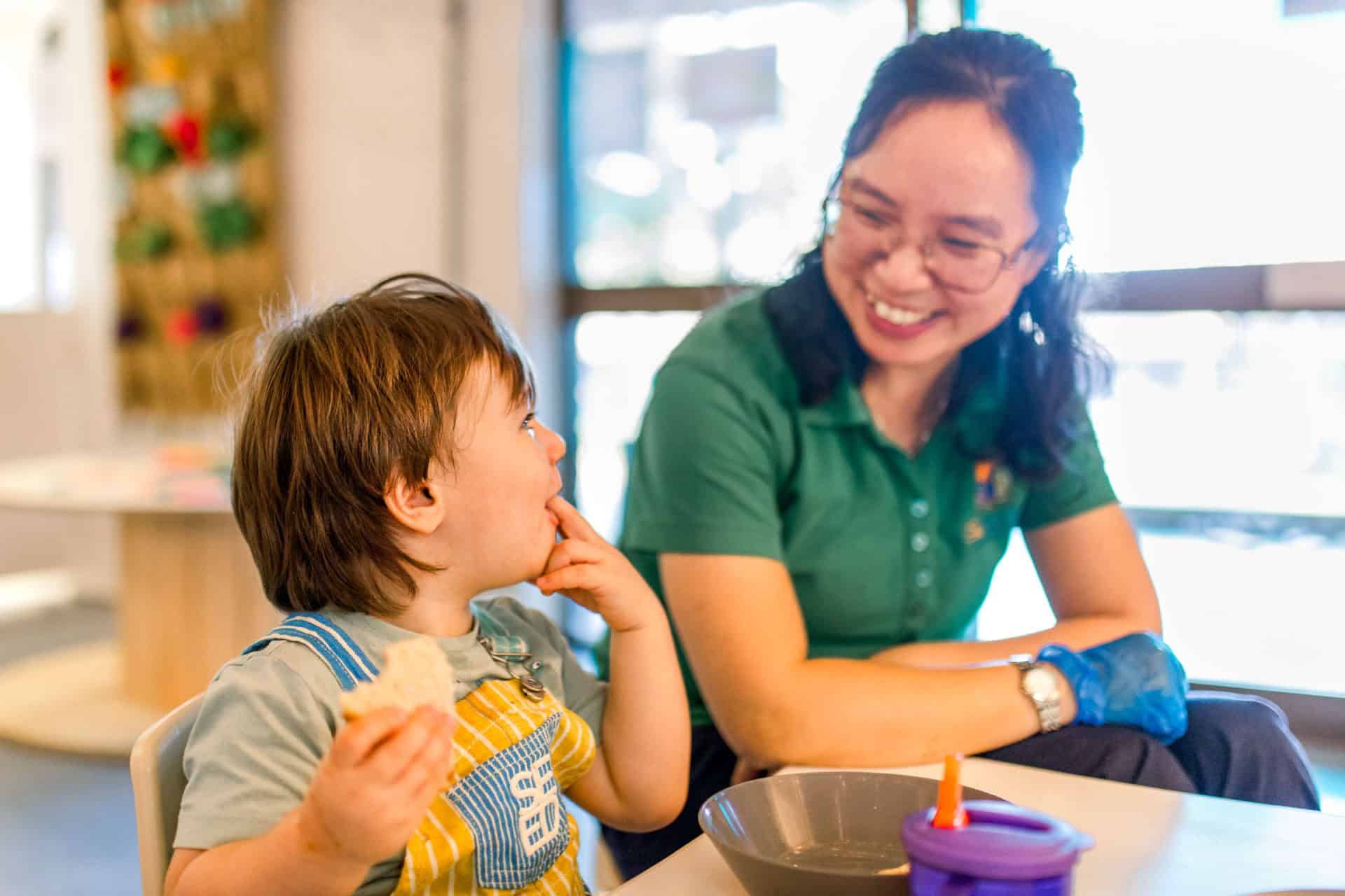 Toddler in a bib sitting at a table eating a snack with a smiling female childcare educator